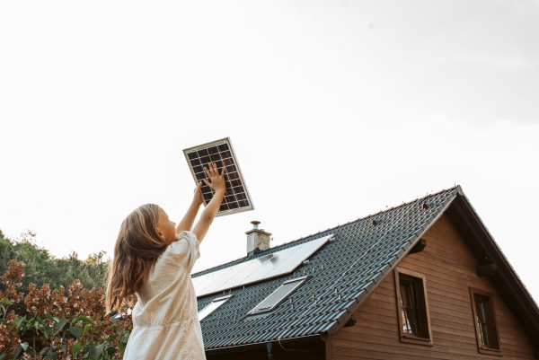 Little girl with model of solar panel, standing in the middle of meadow, house with solar panels behind. Concept of renewable resources, sustainability and green energy.