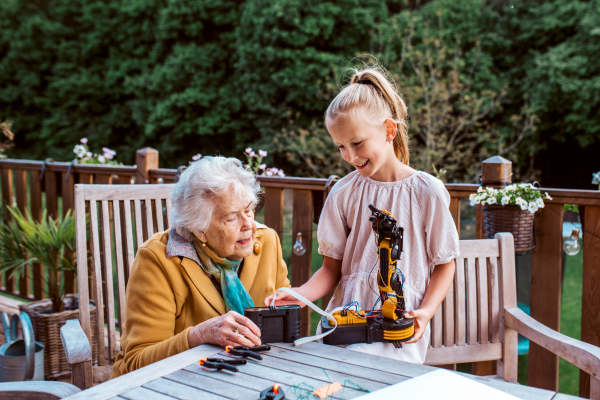 Elderly grandmother supporting granddaughter in robotics, science, IT technologies and programming. Young girl assembling a simple robot and shoving it to grandma.