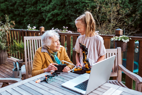 Elderly grandmother supporting granddaughter in robotics, science, IT technologies and programming. Young girl assembling a simple robot and shoving it to grandma.