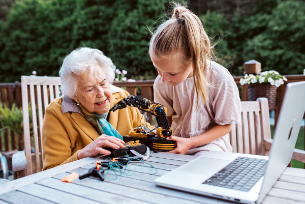 Elderly grandmother supporting granddaughter in robotics, science, IT technologies and programming. Young girl assembling a simple robot and shoving it to grandma.