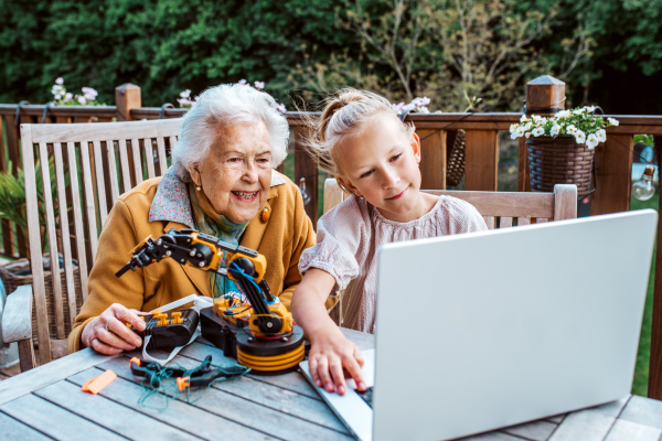 Elderly grandmother supporting granddaughter in robotics, science, IT technologies and programming. Young girl assembling a simple robot and shoving it to grandma.