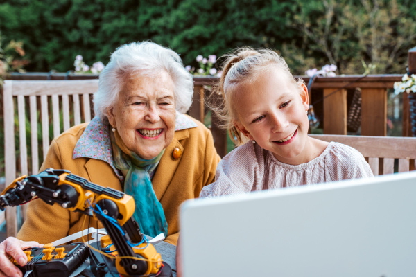 Elderly grandmother supporting granddaughter in robotics, science, IT technologies and programming. Young girl assembling a simple robot and shoving it to grandma.