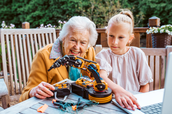 Elderly grandmother supporting granddaughter in robotics, science, IT technologies and programming. Young girl assembling a simple robot and shoving it to grandma.