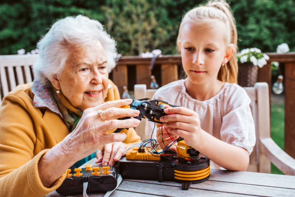 Elderly grandmother supporting granddaughter in robotics, science, IT technologies and programming. Young girl assembling a simple robot and shoving it to grandma.