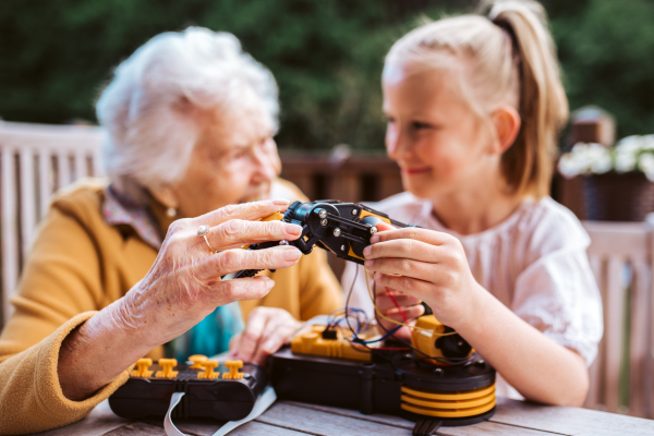 Elderly grandmother supporting granddaughter in robotics, science, IT technologies and programming. Young girl assembling a simple robot and shoving it to grandma.