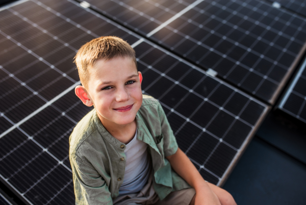 Cute boy sitting on roof with solar panels, looking at camera. Sustainable future for next generation concept.