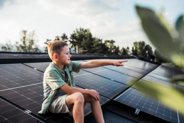 Cute boy sitting on roof with solar panels, looking at surrounding nature. Sustainable future for next generation concept.