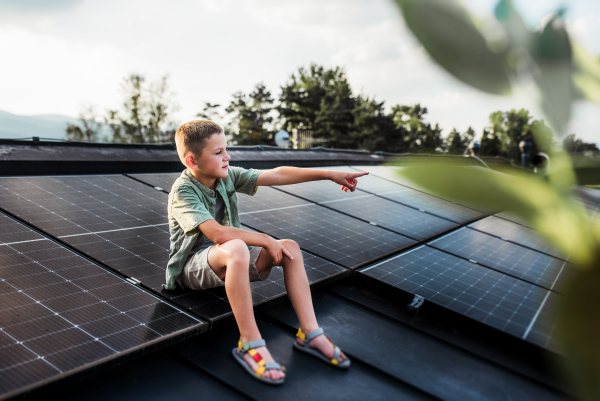 Cute boy sitting on roof with solar panels, looking at surrounding nature. Sustainable future for next generation concept.