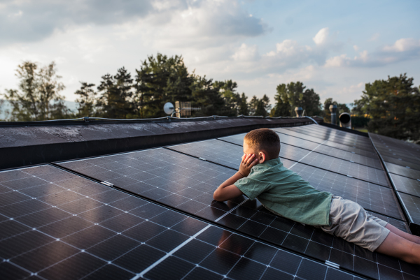 Boy lying on his belly on roof with solar panels, looking at surrounding nature. Sustainable future for next generation concept.