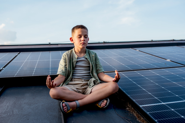 Cute boy sitting on roof with solar panels, meditating with closed eyes. Sustainable future for next generation concept.