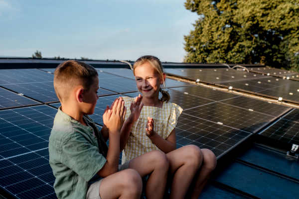 Two young siblings sitting on roof with solar panels, looking at each other.Sustainable future for next generation concept.