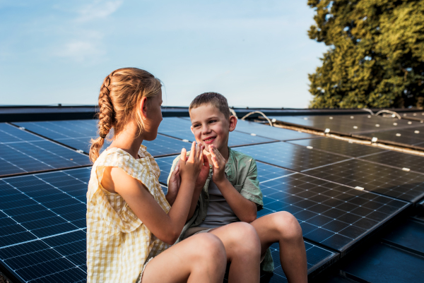 Two young siblings sitting on roof with solar panels, looking at each other.Sustainable future for next generation concept.