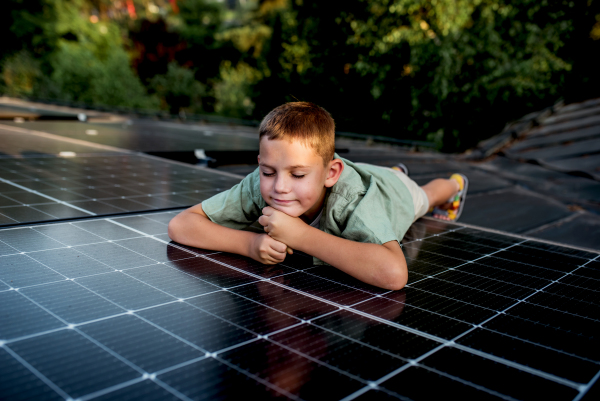 Boy lying on his belly on roof with solar panels, looking at surrounding nature. Sustainable future for next generation concept.