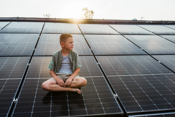 Cute boy sitting on roof with solar panels, looking at surrounding nature. Sustainable future for next generation concept.