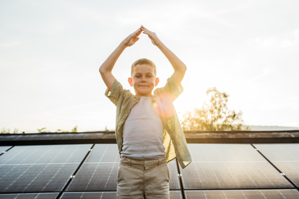 Cute boy making roof gesture with arms, standing on roof with solar panels. Rooftop solar or photovoltaic system. Sustainable future for next generation concept.