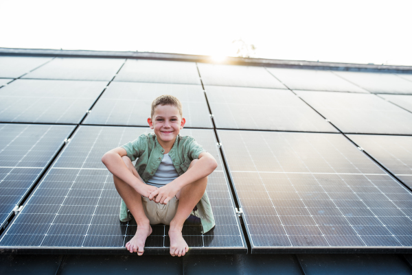 Cute boy sitting on roof with solar panels, looking at camera. Sustainable future for next generation concept.