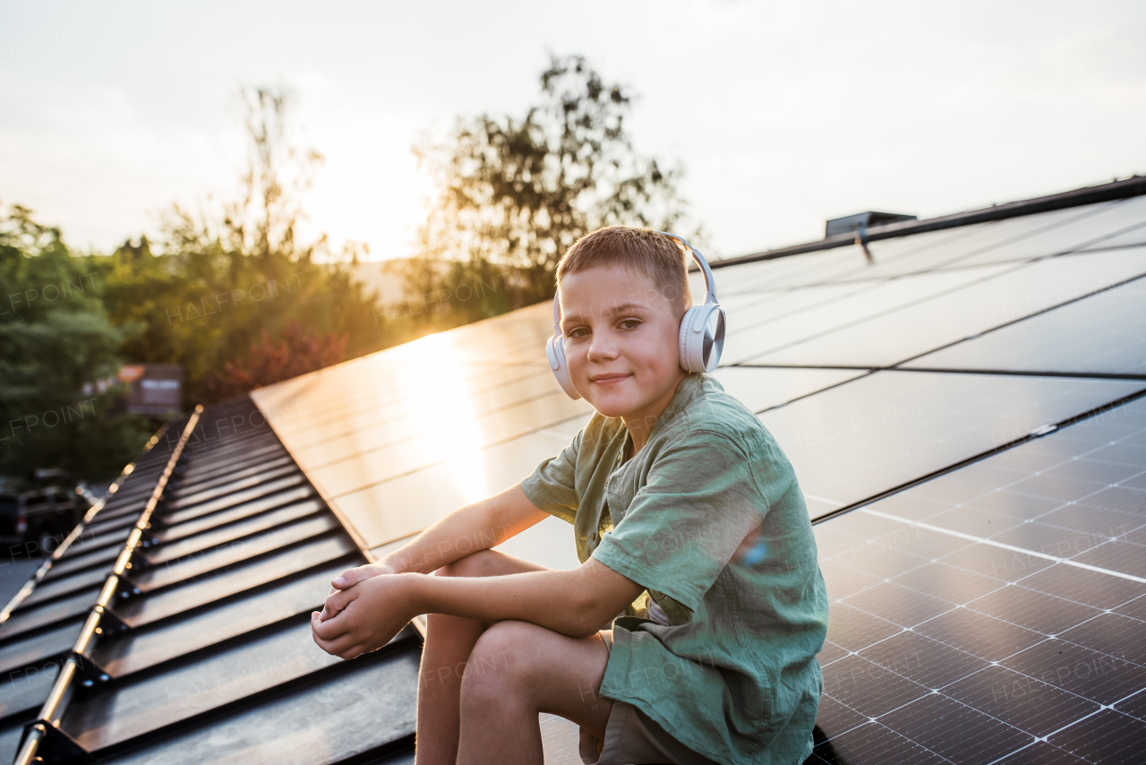 Cute boy sitting on roof with solar panels, headphones on head, listening music. Sustainable future for next generation concept.