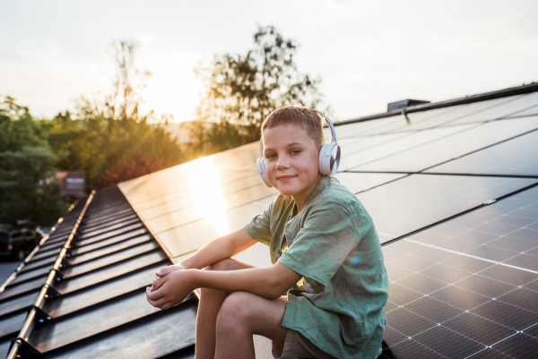 Cute boy sitting on roof with solar panels, headphones on head, listening music. Sustainable future for next generation concept.