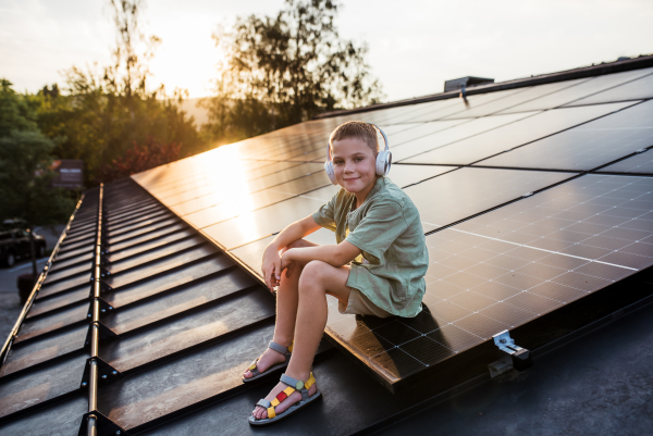 Cute boy sitting on roof with solar panels, headphones on head, listening music. Sustainable future for next generation concept.