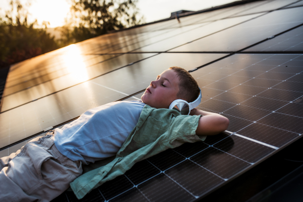 Boy lying on roof with solar panels, eyes closed,headphones on head.. Sustainable future for next generation concept.