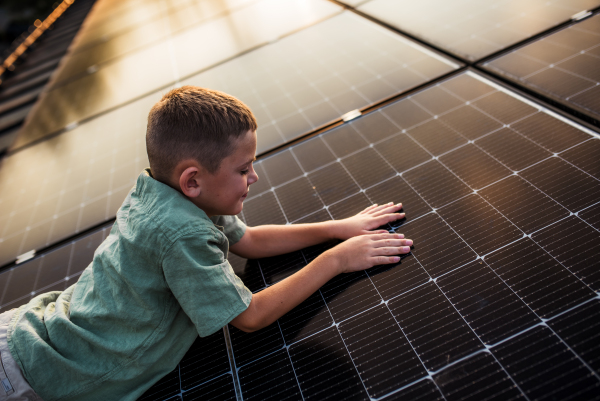 Boy lying on belly on roof with solar panels, touching panel with hands. Sustainable future for next generation concept.