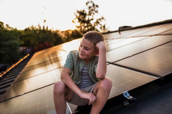 Cute boy sitting on roof with solar panels, looking at surrounding nature. Sustainable future for next generation concept.