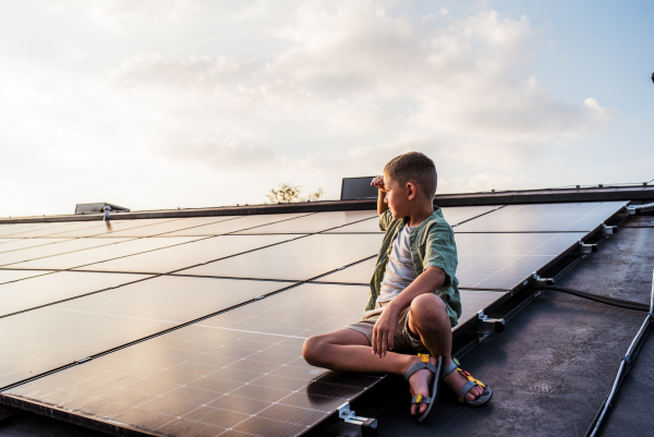 Cute boy sitting on roof with solar panels, looking at sunset. Sustainable future for next generation concept.