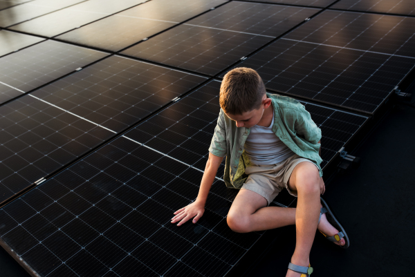 Boy sitting on roof with solar panels, touching panel with hands. Sustainable future for next generation concept.