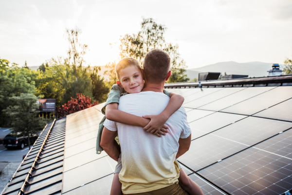 Dad holding girl, standing in front of roof with solar panels during sunset. Rooftop solar or photovoltaic system. Sustainable future for next generation concept.