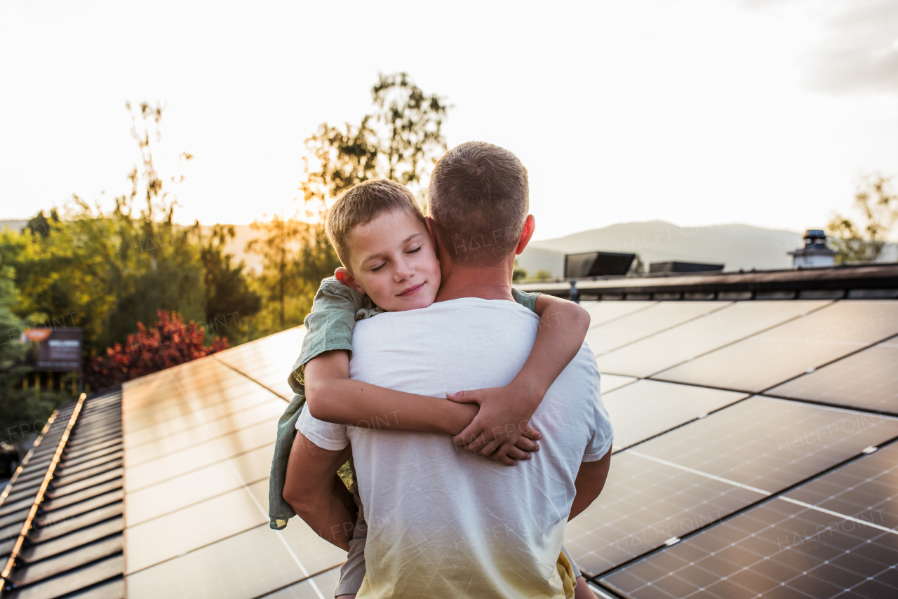 Dad holding boy, standing in front of roof with solar panels during sunset. Rooftop solar or photovoltaic system. Sustainable future for next generation concept.