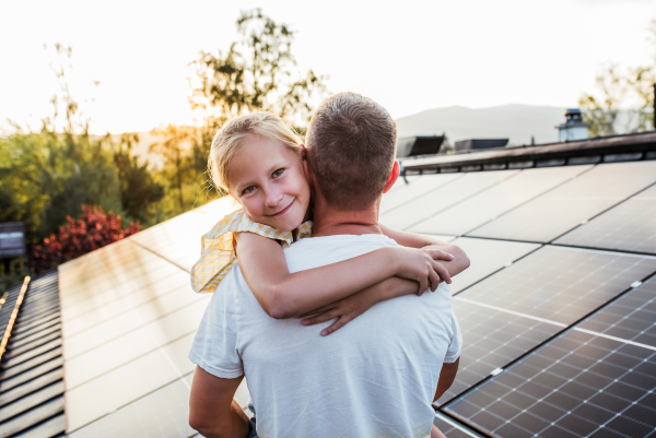 Dad holding girl, standing in front of roof with solar panels during sunset. Rooftop solar or photovoltaic system. Sustainable future for next generation concept.