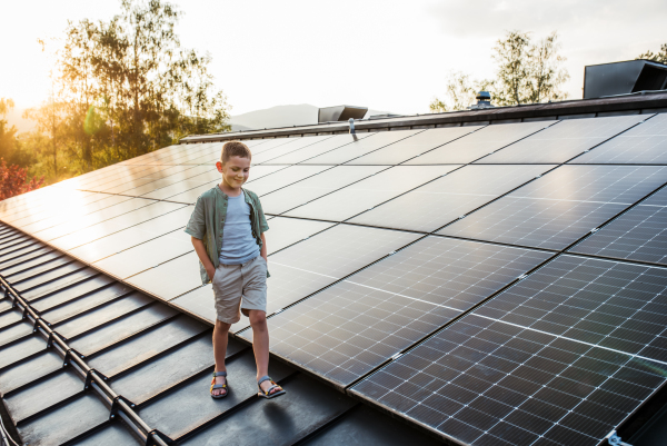 Boy walking on roof with solar panels during sunset, smiling, hands in pockets. Sustainable future for next generation concept.