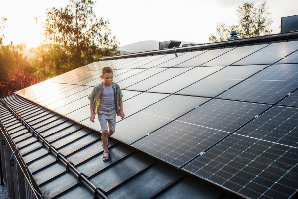 Boy walking on roof with solar panels during sunset, smiling, hands in pockets. Sustainable future for next generation concept.