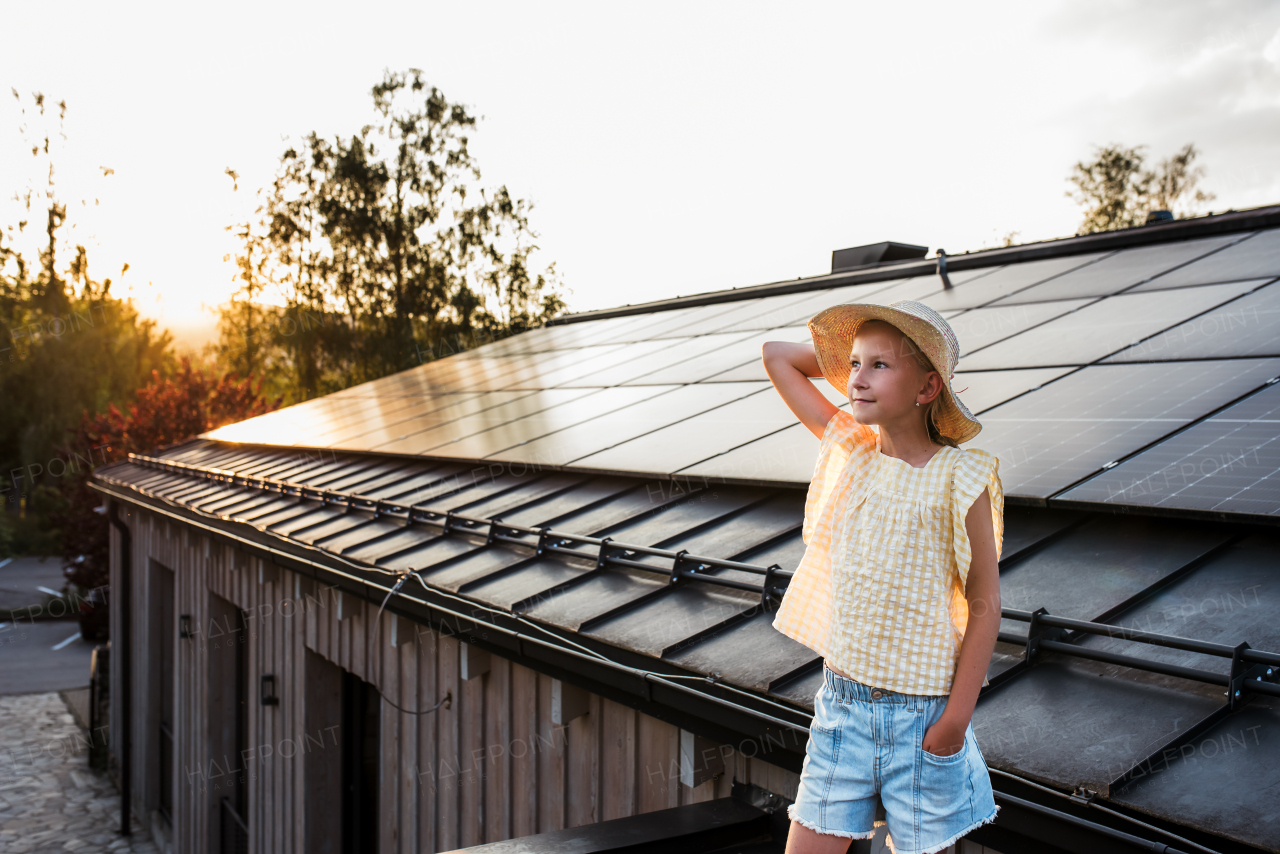 Girl standing in front of roof with solar panels during sunset, smiling and holding her hat. Sustainable future for next generation concept.