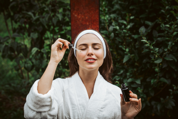 Beautiful woman doing her skin care routine outdoors, wearing bathrobe. Woman using facial skin serum