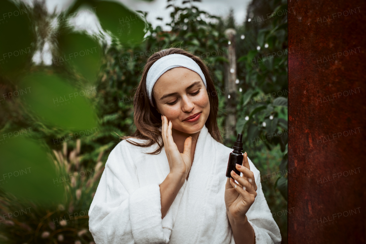 Beautiful woman doing her skin care routine outdoors, wearing bathrobe. Woman using facial skin serum