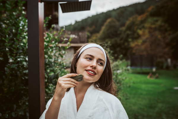 Beautiful woman massaging her face with green jade gua sha tool. Outdoor skin care routine for young woman