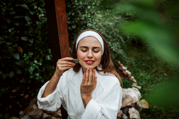 Beautiful woman massaging her face with green jade gua sha tool. Outdoor skin care routine for young woman