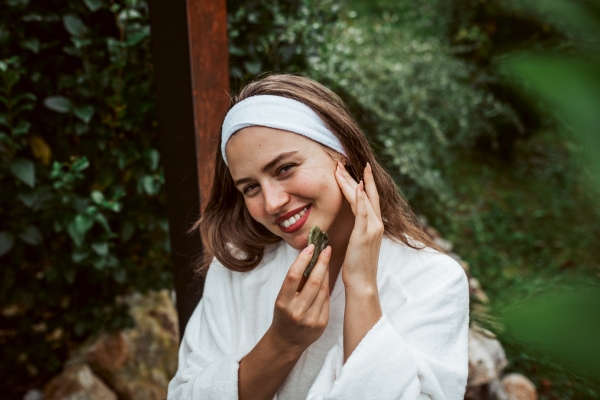 Beautiful woman massaging her face with green jade gua sha tool. Outdoor skin care routine for young woman