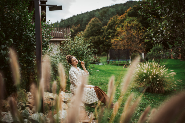 Woman massaging her face with rose quartz roller, sitting in the garden. Outdoor skin care routine for young woman