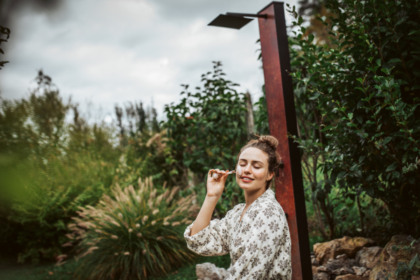 Beautiful woman massaging her face with rose quartz roller, sitting in the garden. Outdoor skin care routine for young woman