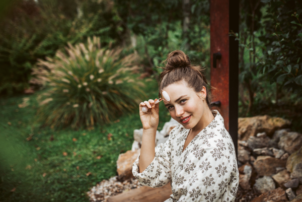 Beautiful woman massaging her face with rose quartz roller, sitting in the garden. Outdoor skin care routine for young woman