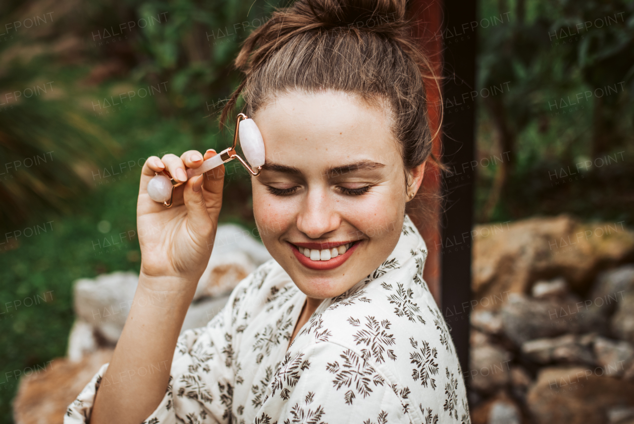 Beautiful woman massaging her face with rose quartz roller, sitting in the garden. Outdoor skin care routine for young woman