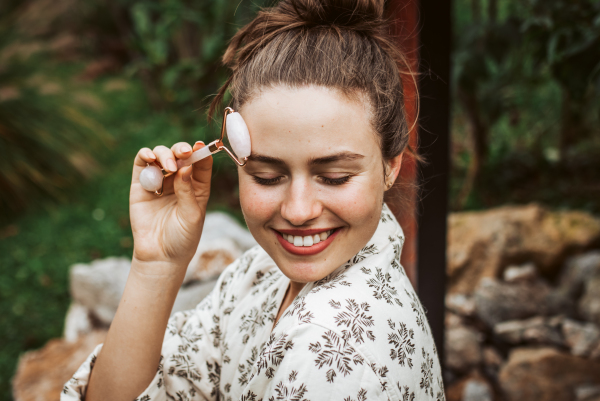 Beautiful woman massaging her face with rose quartz roller, sitting in the garden. Outdoor skin care routine for young woman