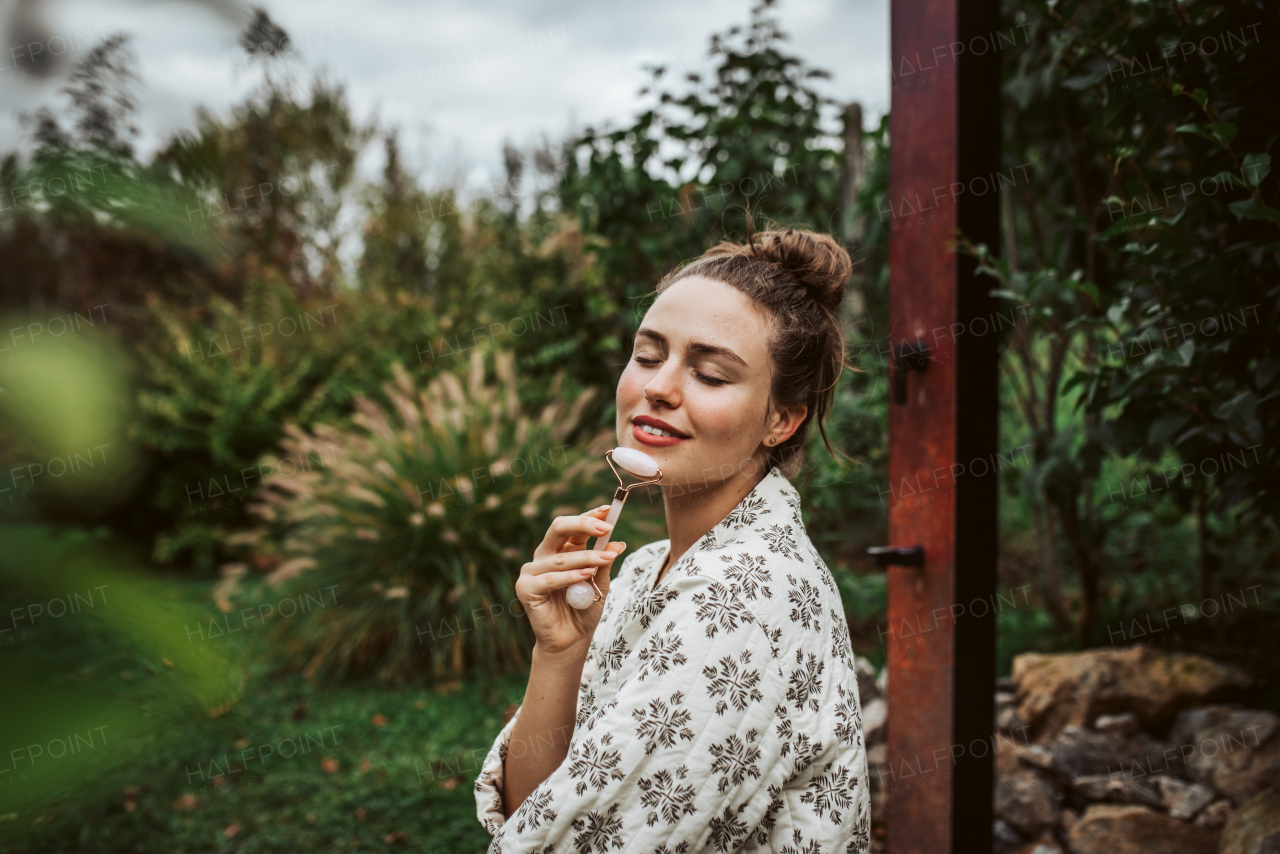 Beautiful woman massaging her face with rose quartz roller, sitting in the garden. Outdoor skin care routine for young woman