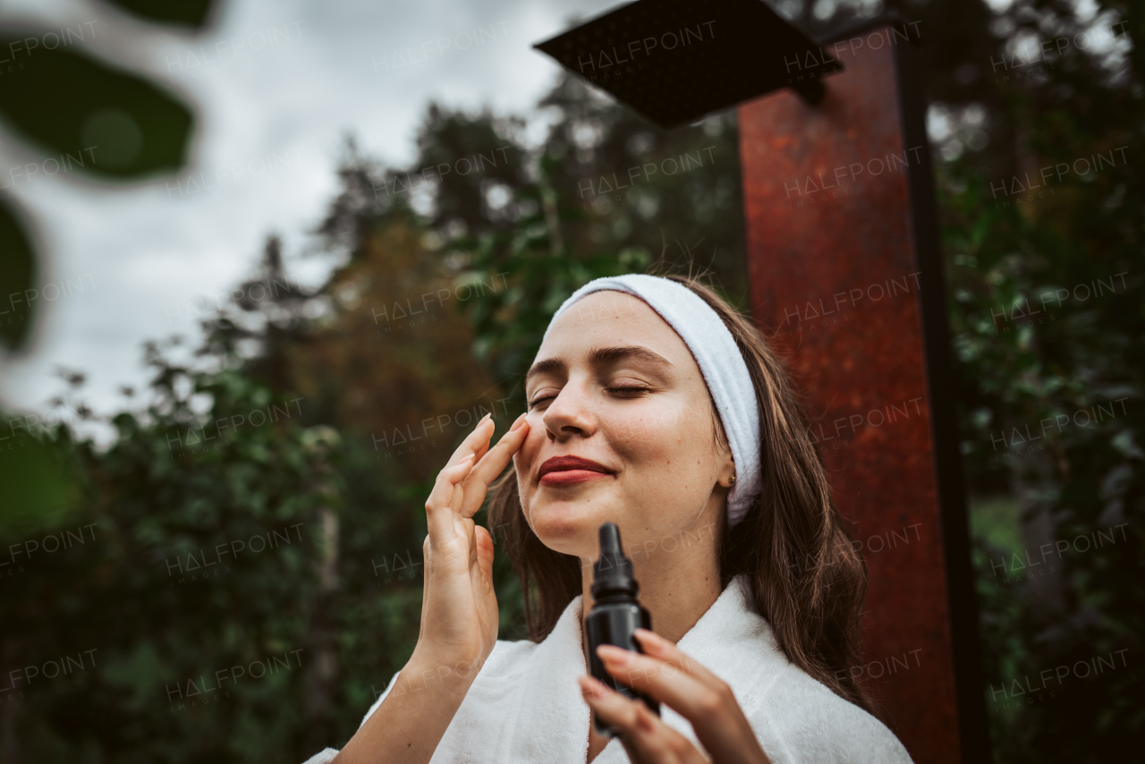 Beautiful woman doing her skin care routine outdoors, wearing bathrobe. Woman using facial skin serum