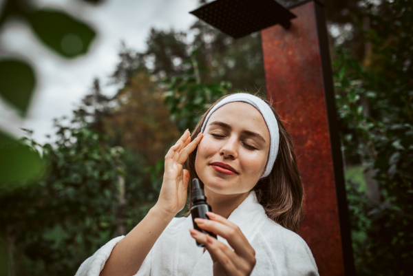 Beautiful woman doing her skin care routine outdoors, wearing bathrobe. Woman using facial skin serum