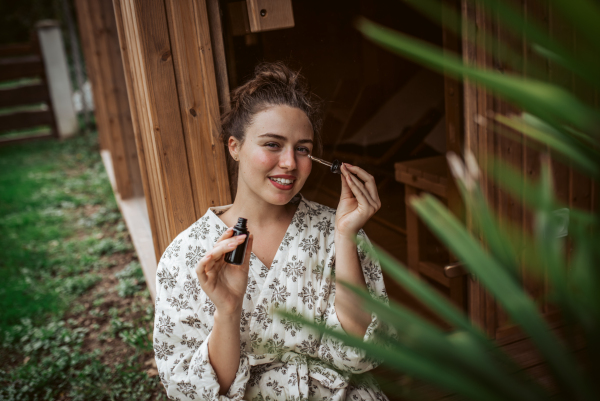 Beautiful woman doing her skin care routine outdoors, wearing bathrobe. Woman using facial skin serum