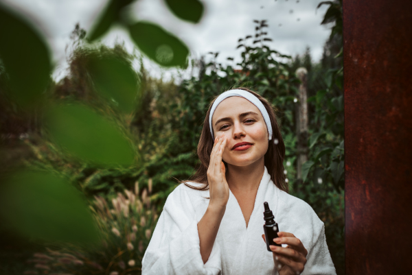 Beautiful woman doing her skin care routine outdoors, wearing bathrobe. Woman using facial skin serum