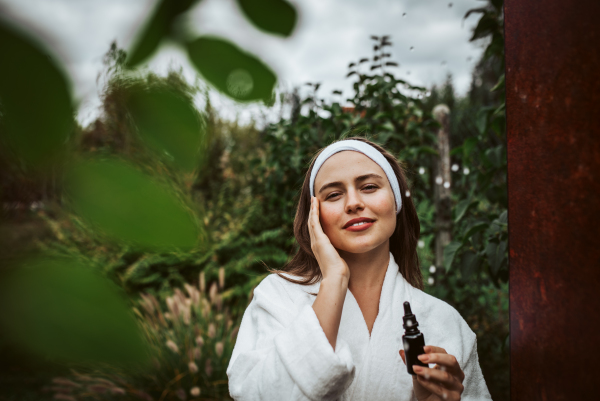 Beautiful woman doing her skin care routine outdoors, wearing bathrobe. Woman using facial skin serum